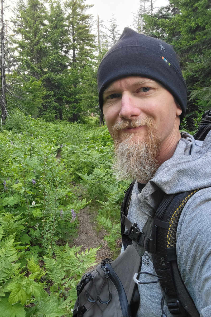 Erick looking into the camera along a trail with lush vegetation and trees in the background